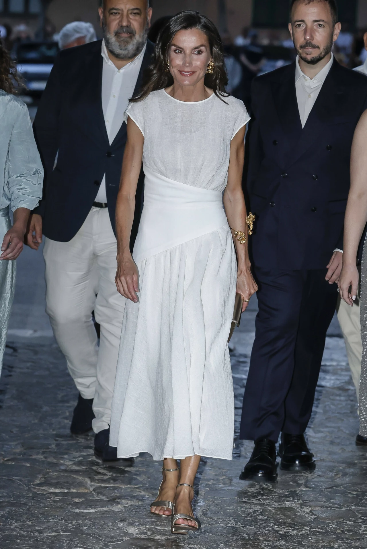 Królowa Letizia wybrała niezwykle prostą, ale elegancką kreację Elegancka kobieta w białej sukience idąca po kamiennej nawierzchni, otoczona przez kilku mężczyzn ubranych w garnitury oraz inną osobę w jasnoniebieskim stroju, wszyscy idą razem wieczorową porą.