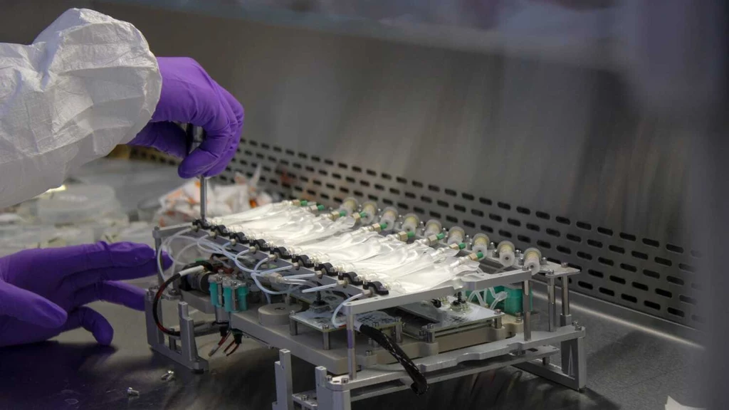 Ręka w rękawiczce laboratoryjnej pracuje nad specjalistycznym urządzeniem z wieloma przewodami i podzespołami elektronicznymi, umieszczonym na metalowym blacie pod wentylowaną osłoną.