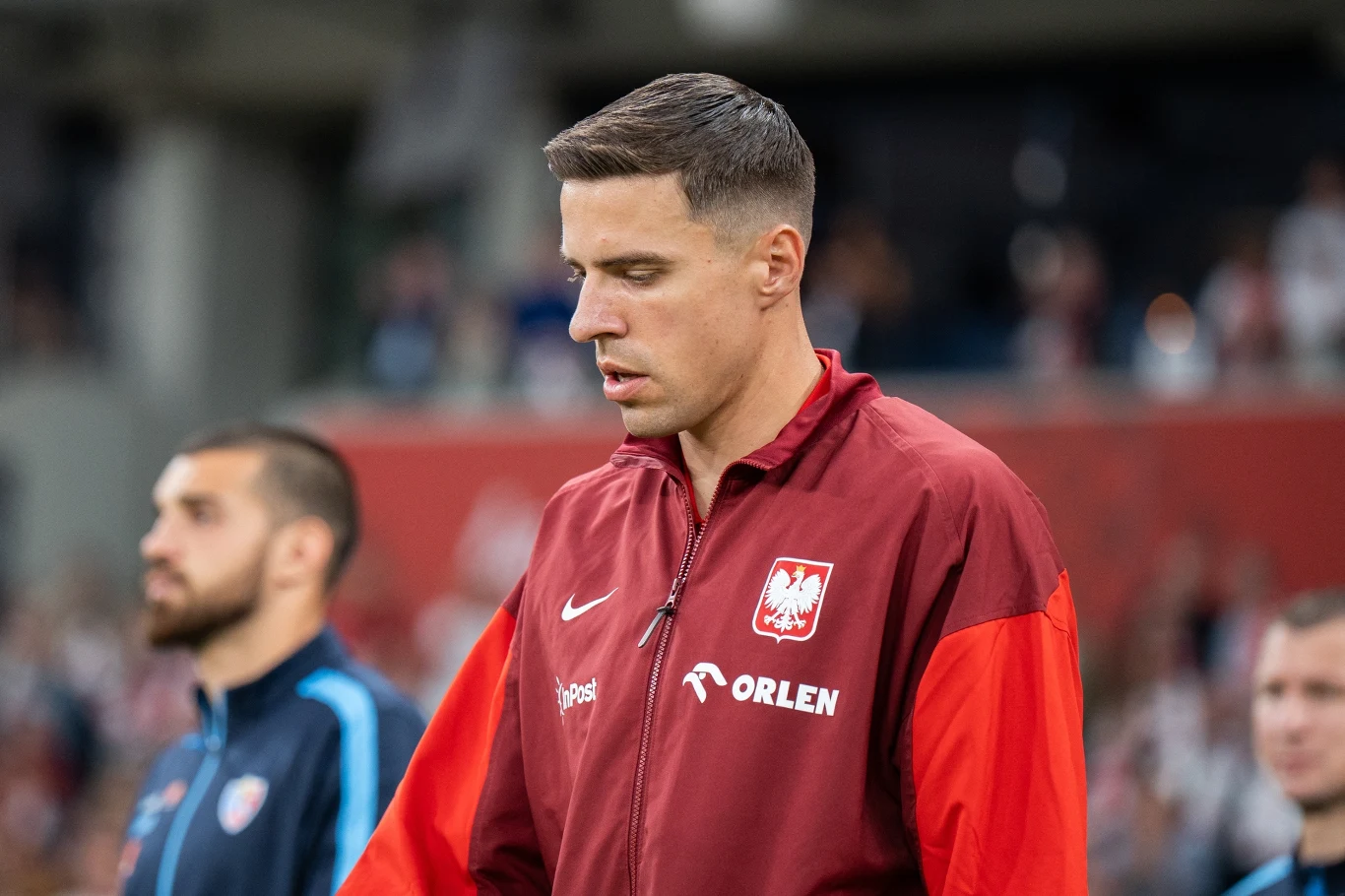 Piłkarz w czerwonej reprezentacyjnej kurtce Polski z herbem orła i logiem sponsora, stoi na tle rozmytego stadionu wśród innych sportowców.