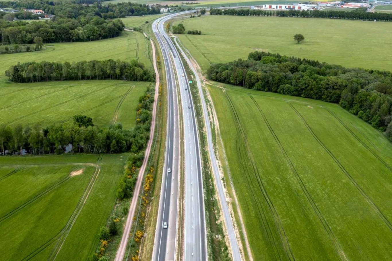Nowoczesna droga szybkiego ruchu prowadząca przez zielone pola i lasy, otoczona pasami roślinności oraz polami uprawnymi, kilka samochodów jedzie w obu kierunkach.