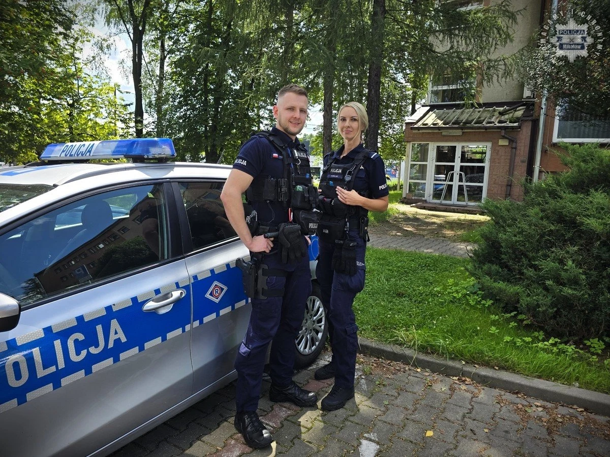 Sierż. Dawid Furman oraz st. post. Patrycja Wolny po otrzymaniu zgłoszenia natychmiast udali się we wskazane miejsce. Dwóch policjantów w mundurach stoi obok radiowozu zaparkowanego na chodniku, w tle widoczny budynek i zieleń.