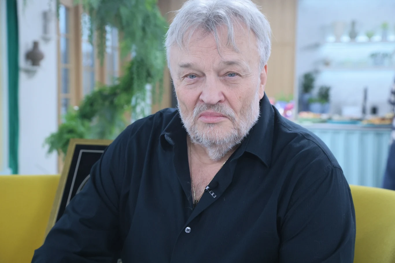 Krzysztof Cugowski komentuje decyzję syna Starszy mężczyzna z siwymi włosami i brodą, ubrany w czarną koszulę, siedzi na żółtej sofie w jasnym, nowoczesnym wnętrzu z zielenią w tle.