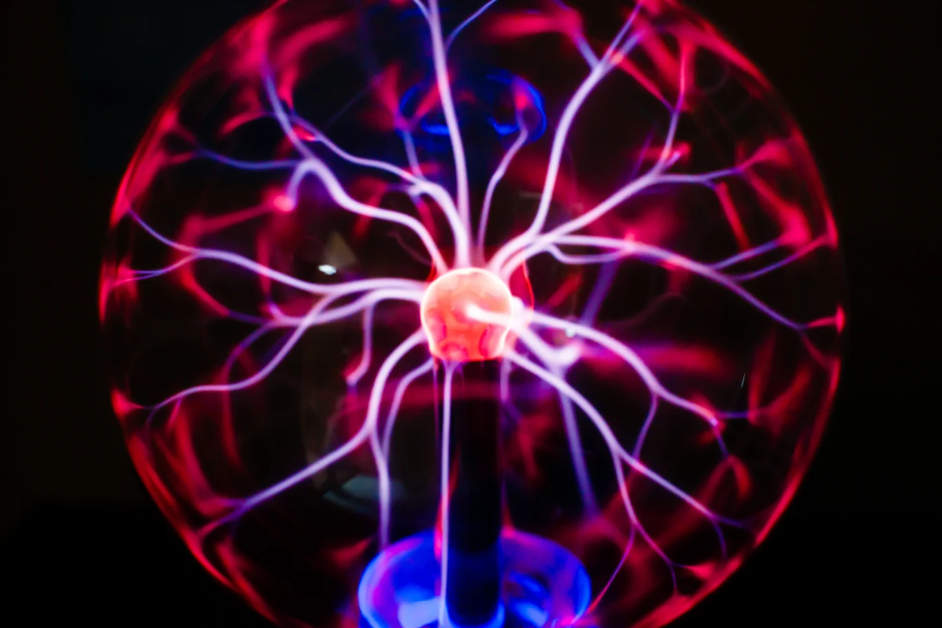 Plazma to zjonizowany gaz tj. naładowane w niej cząstki elektronów i jonów poruszają się swobodnie, tworząc unikalne właściwości, takie jak przewodnictwo elektryczne. Plazma pyłowa dodaje naładowane cząstki pyłu Szklana kula plazmowa z centralną elektroda emitującą kolorowe, rozgałęzione wyładowania elektryczne w odcieniach różu, niebieskiego i fioletu, rozchodzące się w kierunku szkła.