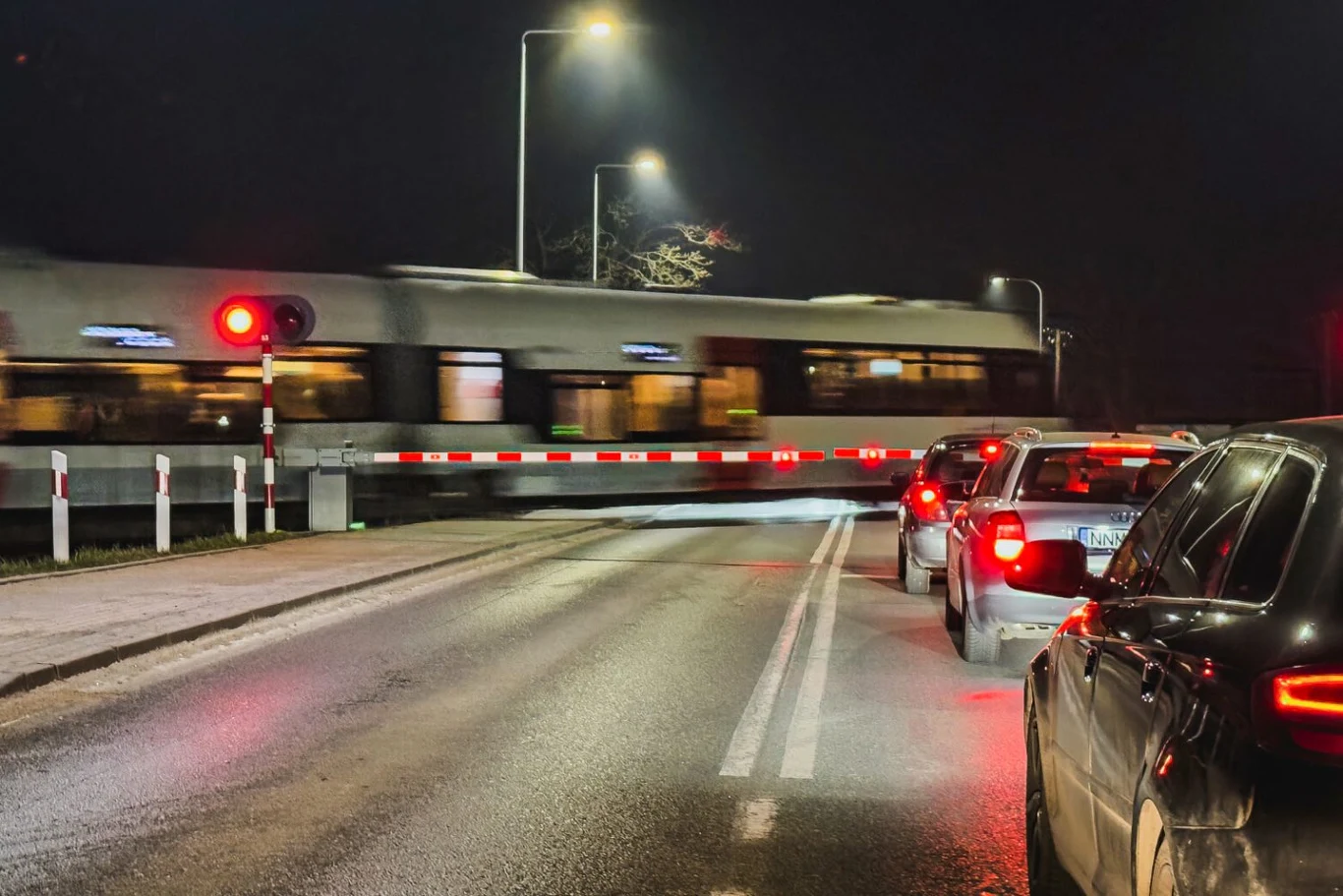 Chwila nieuwagi mogła kosztować życie. Policjanci uratowali dziecko na torach. / zdj. ilustracyjne Szlaban na przejeździe kolejowym zamknięty po zmroku, z przejeżdżającym pociągiem pasażerskim, przed którym stoi rząd czekających samochodów, widoczne światła czerwone sygnalizujące zakaz wjazdu.