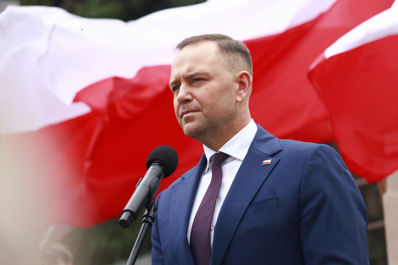 Mężczyzna w garniturze przemawiający do mikrofonu, w tle duża polska flaga, uroczysta atmosfera i powaga sytuacji.