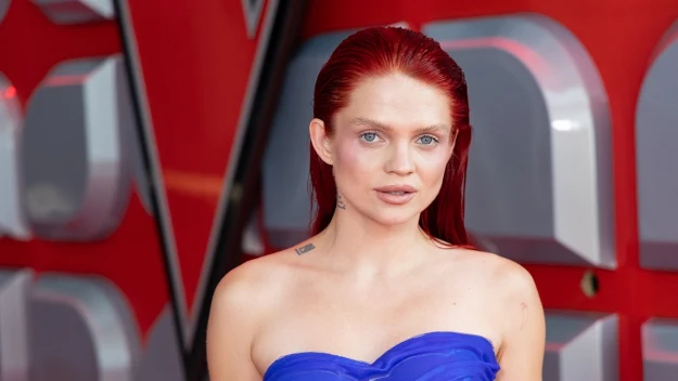 Kobieta o rudych włosach, ubrana w niebieską suknię bez ramiączek, stoi na tle czerwonej ściany z futurystycznymi wzorami, patrząc prosto w obiektyw.