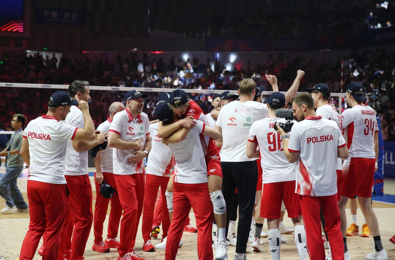 Polscy siatkarze w białych koszulkach i czerwonych spodniach świętują zwycięstwo na parkiecie, obejmując się, podnosząc ręce do góry i okazując radość po zakończonym meczu finałowym w hali sportowej.