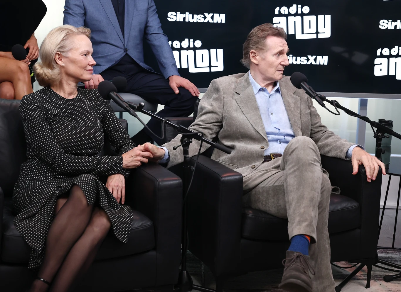 Pamela Anderson i Liam Neeson już nie chcą ukrywać swojego związku Dwoje elegancko ubranych dorosłych siedzi na czarnych fotelach, trzymając się za ręce, rozmawiają ze sobą w studiu radiowym, dookoła widoczne mikrofony i tło z logo SiriusXM oraz radio Andy.