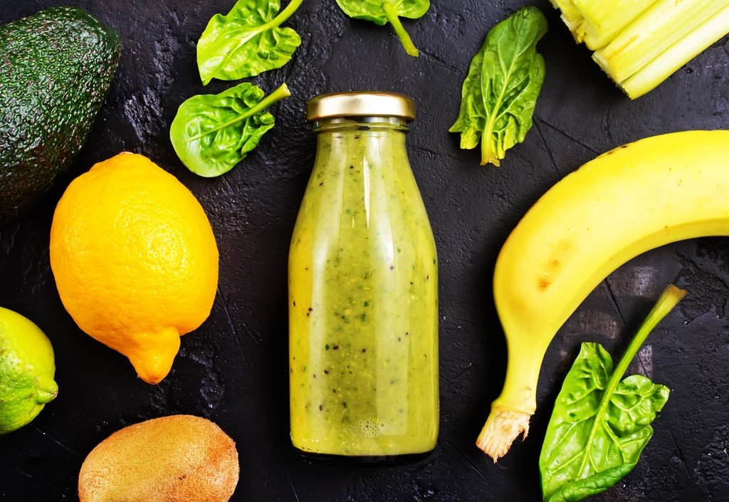 Butelka zielonego smoothie w otoczeniu świeżych owoców i warzyw takich jak banan, cytryna, kiwi, awokado oraz liście szpinaku.