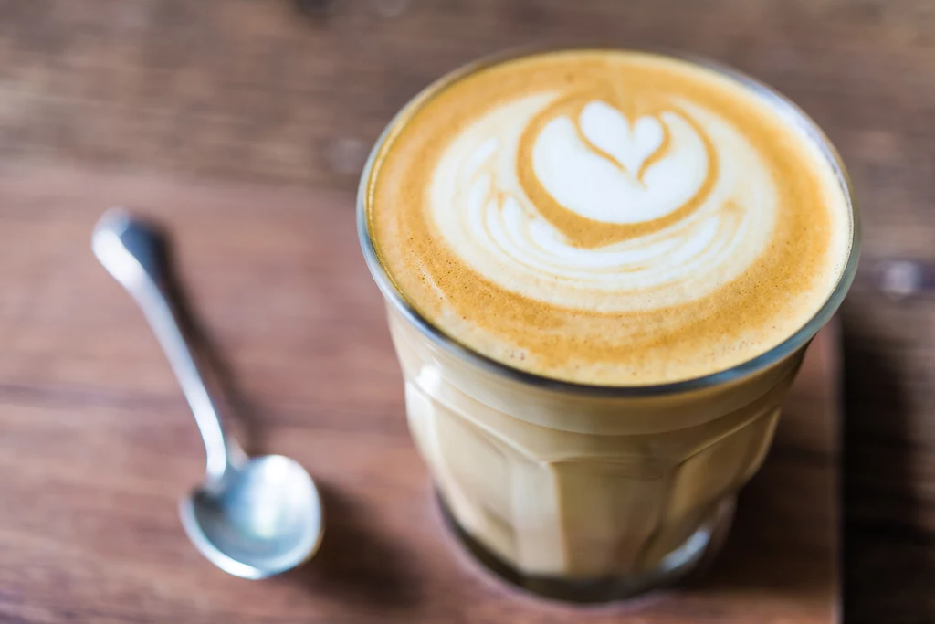 Kawa latte w szklance z artystycznym wzorem z mleka w kształcie serca na piance, obok znajduje się łyżeczka na drewnianej powierzchni.