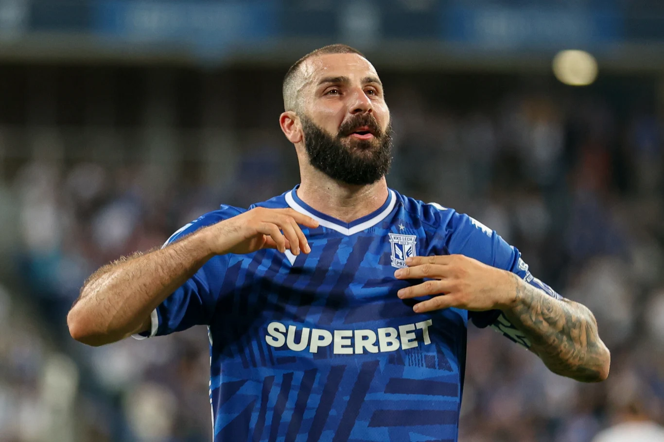 Mikael Ishak, 22.07.2025 r. Piłkarz w niebieskiej koszulce drużyny Lech Poznań, z widocznym logotypem sponsora, wykonuje gesty rękoma na tle rozmytej publiczności na stadionie.