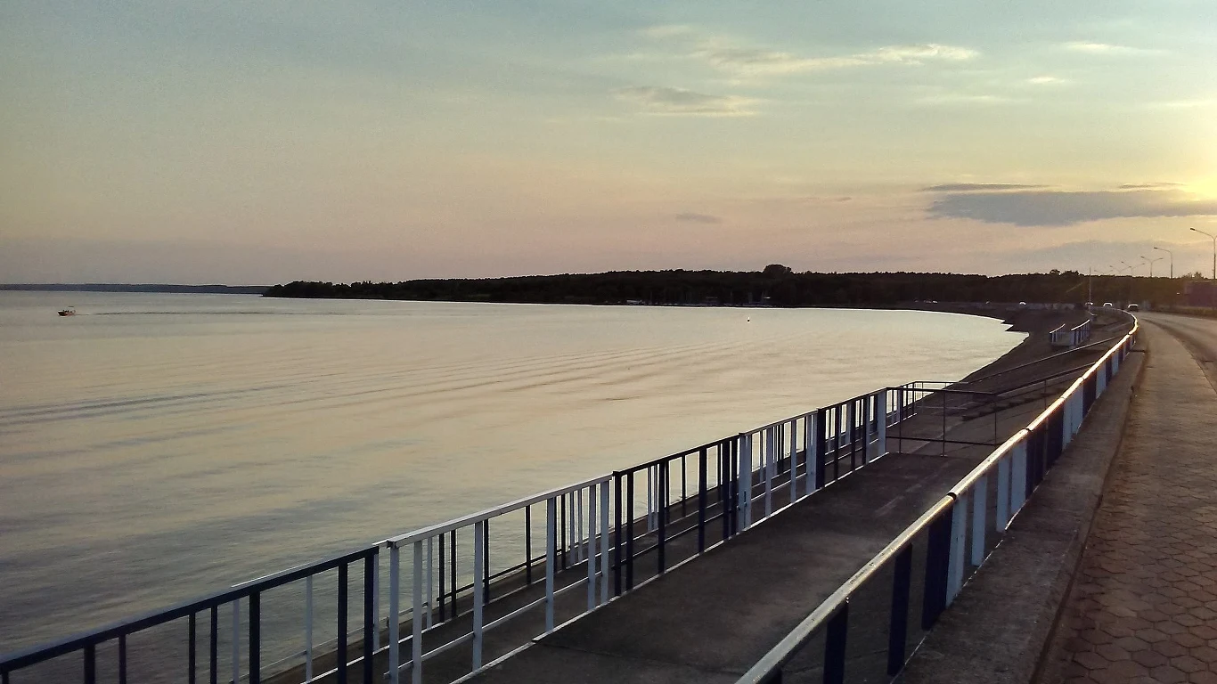 Gigantyczna rewitalizacja Zalewu Sulejowskiego ruszy już wkrótce Nadbrzeżna promenada z metalową barierką rozciąga się wzdłuż spokojnej tafli jeziora, na drugim planie widoczny brzeg porośnięty drzewami, a całość otula łagodne światło zachodzącego słońca.