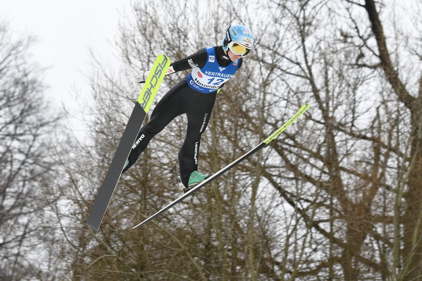 Zawodnik w stroju sportowym podczas skoku narciarskiego unosi się w powietrzu, w tle bezlistne drzewa i zimowa aura.