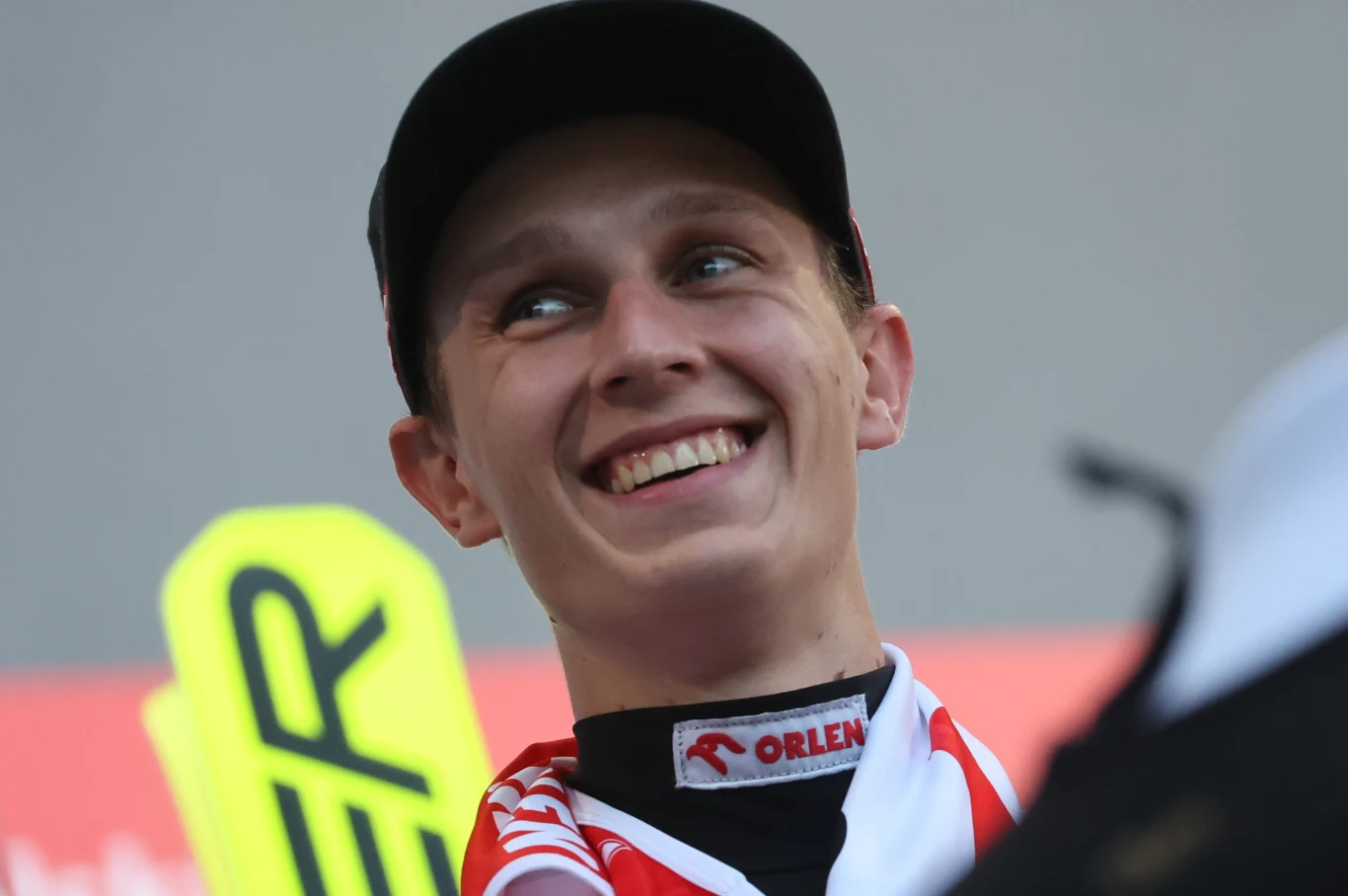 Młody mężczyzna w czapce z daszkiem uśmiecha się szeroko, ubrany w sportową odzież, w tle żółty znak z czarnym napisem oraz czerwone tło.