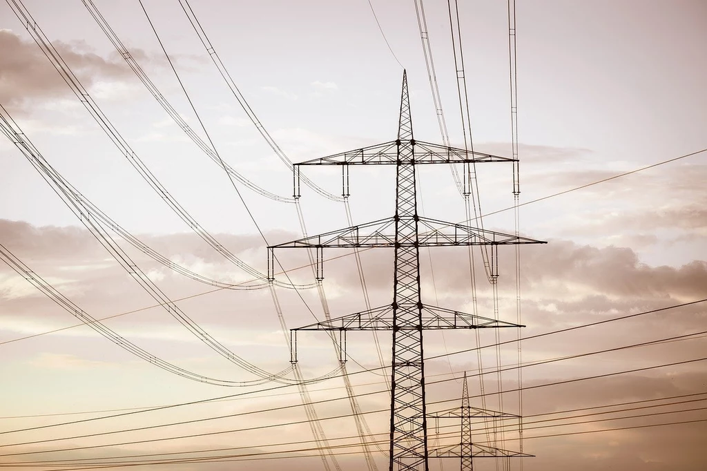 Wieże przesyłowe linii wysokiego napięcia z licznymi przewodami elektrycznymi biegnącymi przez otwartą przestrzeń pod zachmurzonym niebem, dominujące elementy konstrukcyjne stalowe.