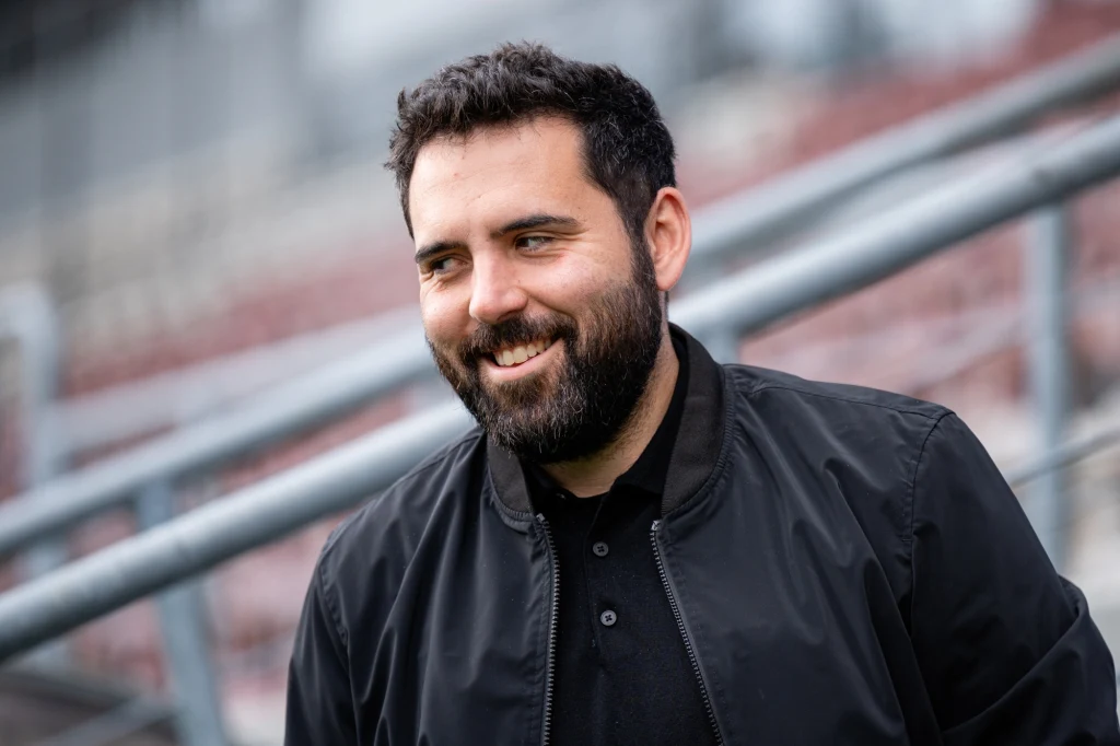 Goncalo Feio Mężczyzna z brodą w ciemnej kurtce uśmiecha się i stoi na tle metalowej konstrukcji, prawdopodobnie na stadionie lub w przestrzeni publicznej.