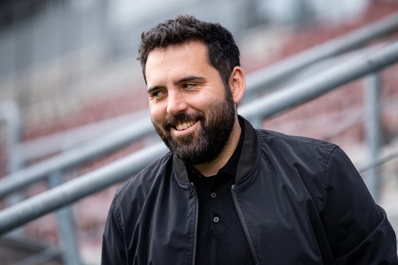 Mężczyzna z brodą w ciemnej kurtce uśmiecha się i stoi na tle metalowej konstrukcji, prawdopodobnie na stadionie lub w przestrzeni publicznej.