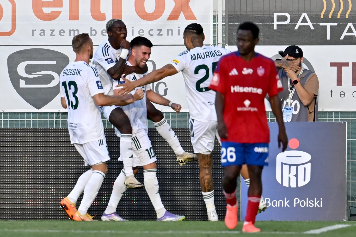 Grupa piłkarzy w białych strojach cieszy się po zdobyciu gola, obejmując się w pobliżu bocznej linii boiska, na pierwszym planie zawodnik w czerwonej koszulce odchodzi z wyrazem rozczarowania na twarzy.