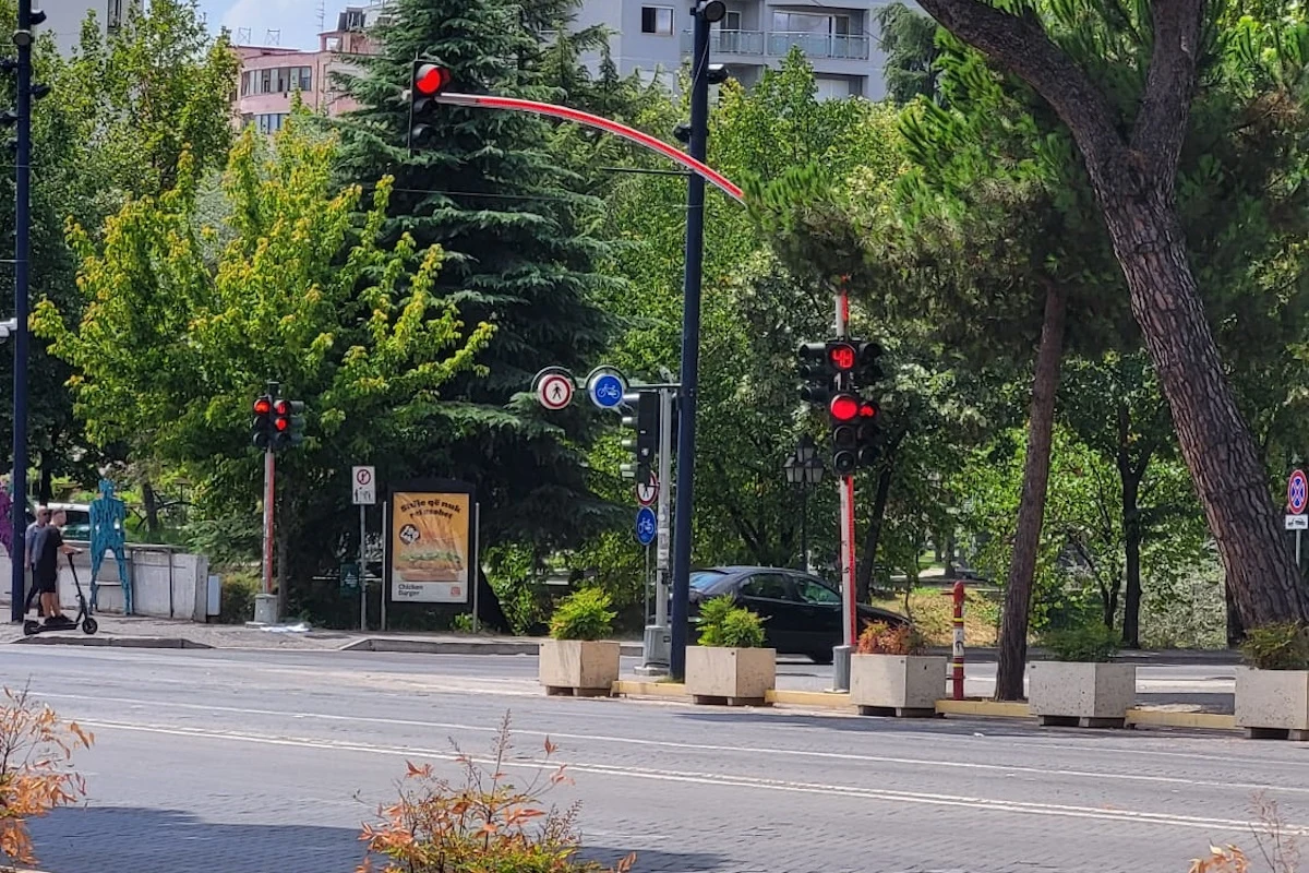 Nietypowe światła w Albanii. Całym słupek jest sygnalizatorem Sygnalizacja świetlna z wyświetlaczem odliczającym czas do zmiany świateł, puste przejście dla pieszych, kilka osób na hulajnogach elektrycznych, duże drzewa oraz zieleń miejska pośród budynków mieszkalnych w tle.