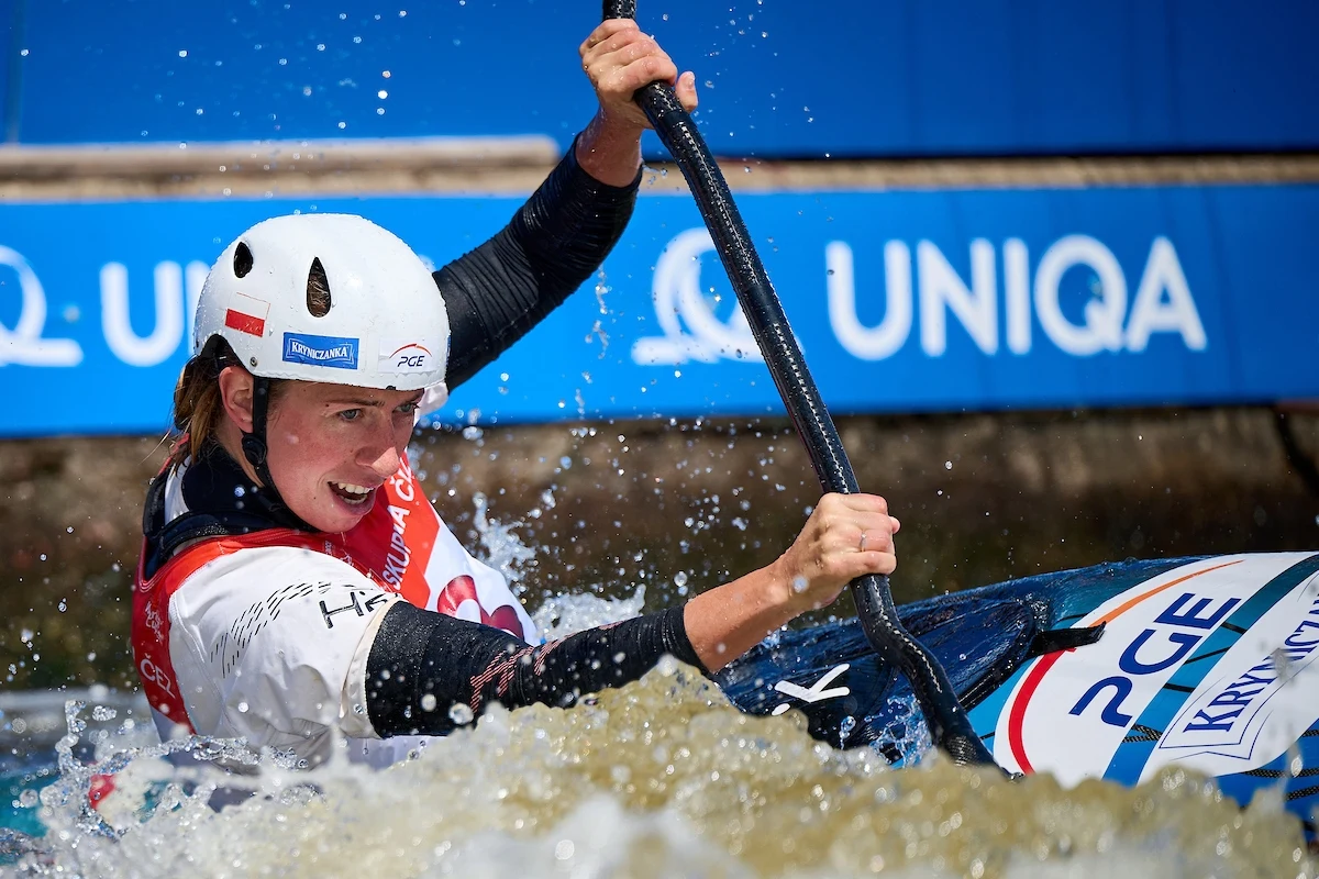 Zawodniczka w kasku i kamizelce ratunkowej intensywnie wiosłuje podczas wyścigu kajakowego na torze wodnym, z dynamicznie rozpryskującą się wodą wokół łódki, na tle baneru z logo sponsora.