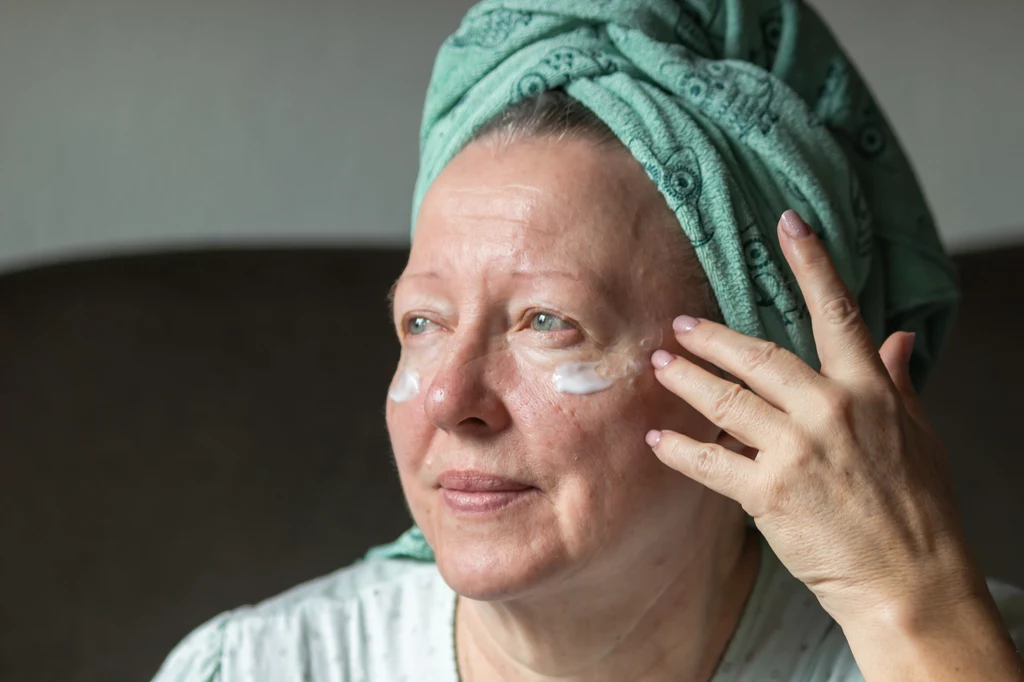 Niektórzy przywiązują się do jednego zestawu kosmetyków, z którego korzystali przez lata Starsza kobieta z zielonym turbanem na głowie nakłada krem pod oczy, delikatnie wklepując produkt w skórę wokół oczu. Na jej twarzy widoczne są zmarszczki świadczące o dojrzałości, a całości towarzyszy spokojny, domowy nastrój.