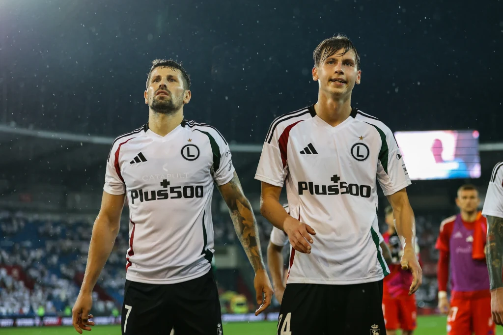 Paweł Wszołek i Jan Ziółkowski Dwóch piłkarzy w białych koszulkach z logo Legii Warszawa wychodzi z boiska podczas meczu, w tle widoczni są inni zawodnicy oraz stadion pełen kibiców.