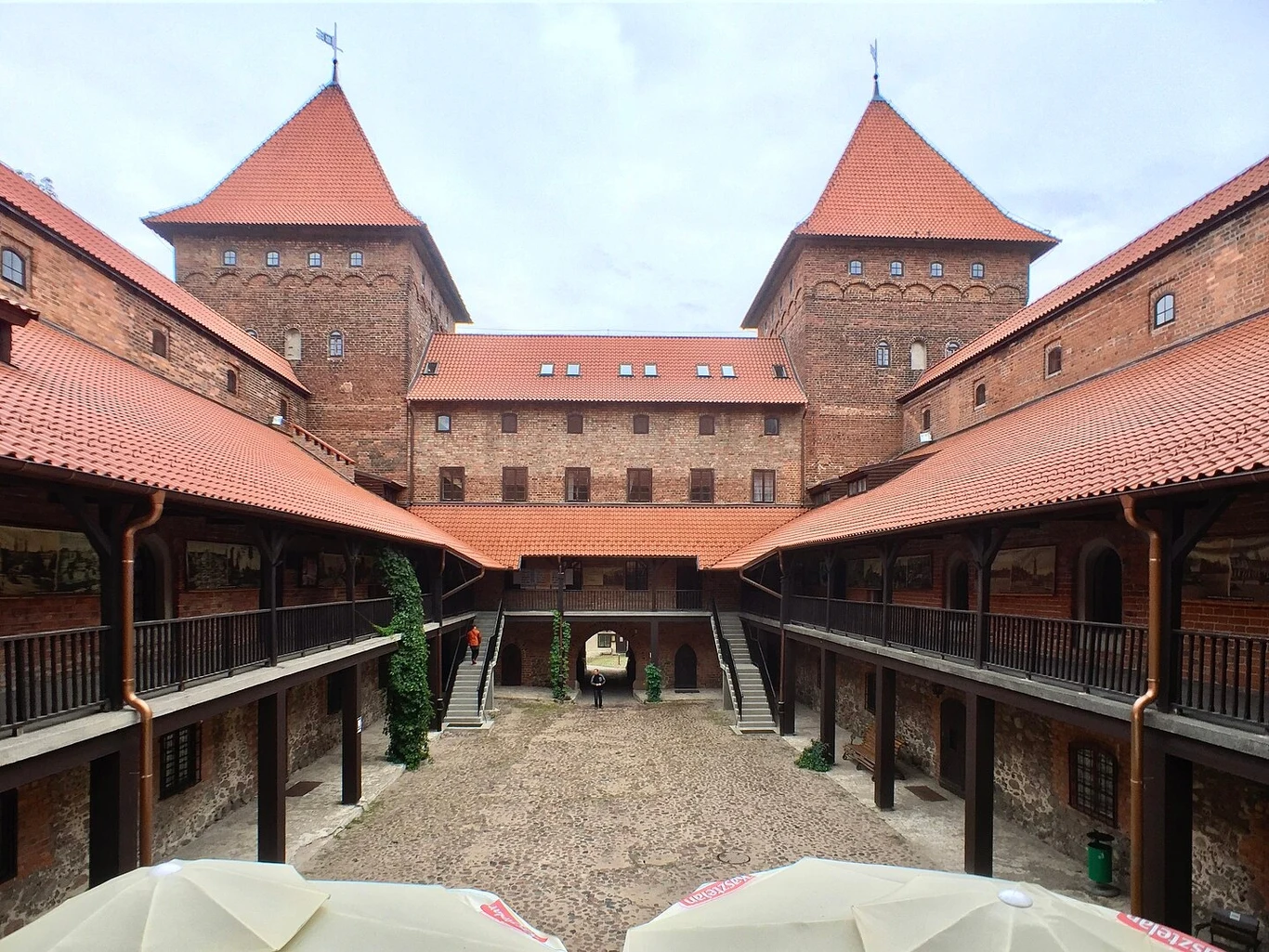 Dziedziniec zamku otoczony ceglanymi murami z arkadami na dwóch poziomach oraz trzema wieżami z czerwonymi dachami, pośrodku brukowane podwórze i samochód dostawczy.