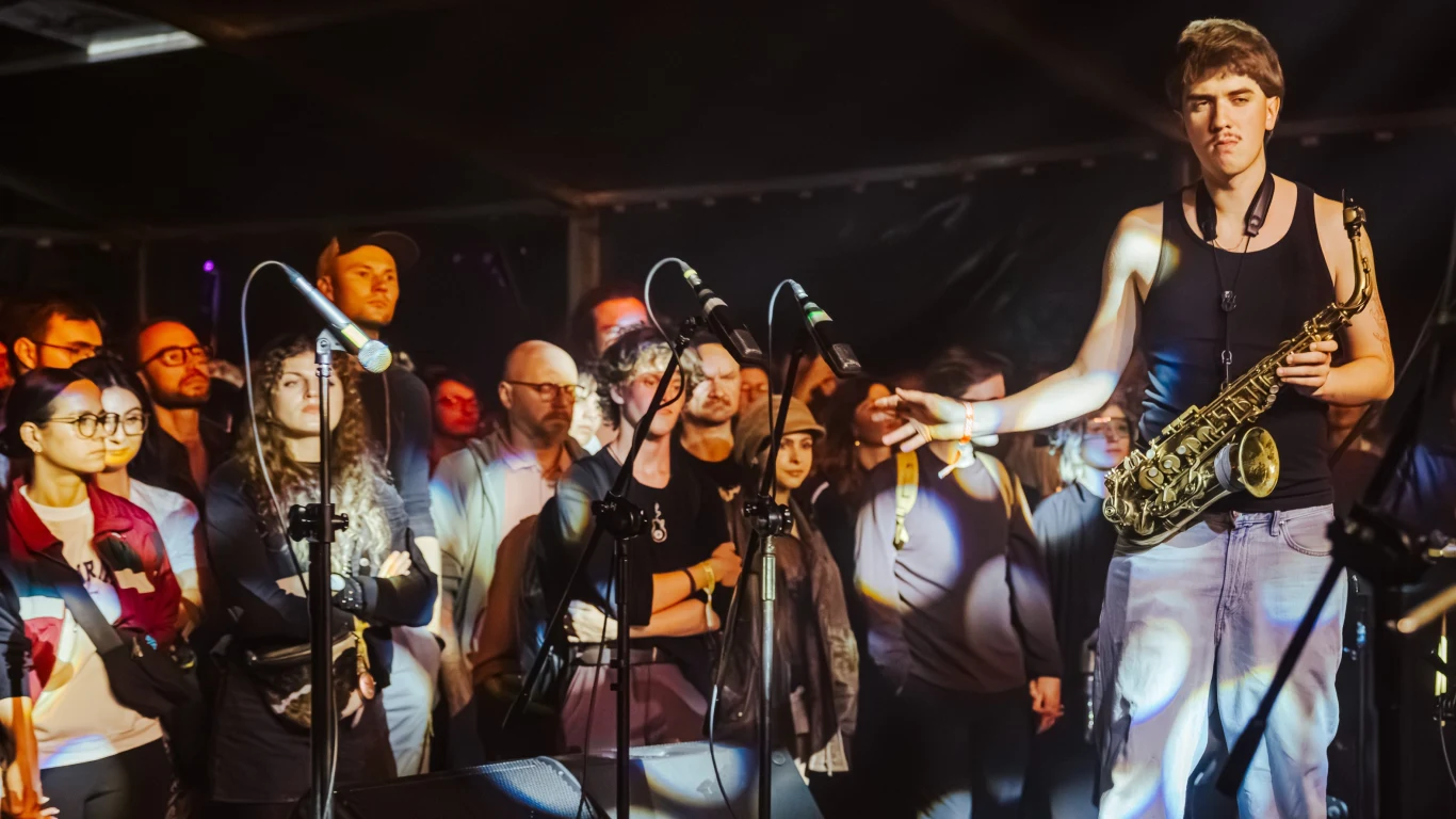 Kosmonauci na OFF Festival Muzyk trzymający saksofon na tle skupionej publiczności, oświetlony scenicznym światłem, wokół widoczne mikrofony i osoby uważnie obserwujące występ.