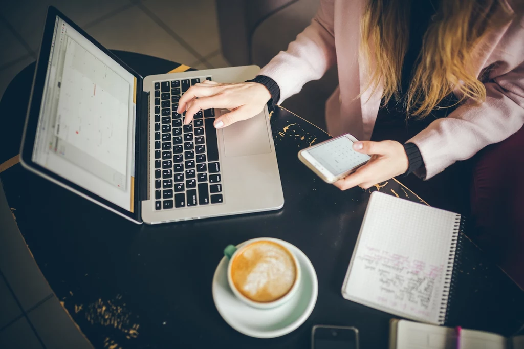 Osoba pracująca przy laptopie, trzymająca telefon w jednej ręce, obok leży notes z zapiskami oraz filiżanka kawy na okrągłym stoliku.
