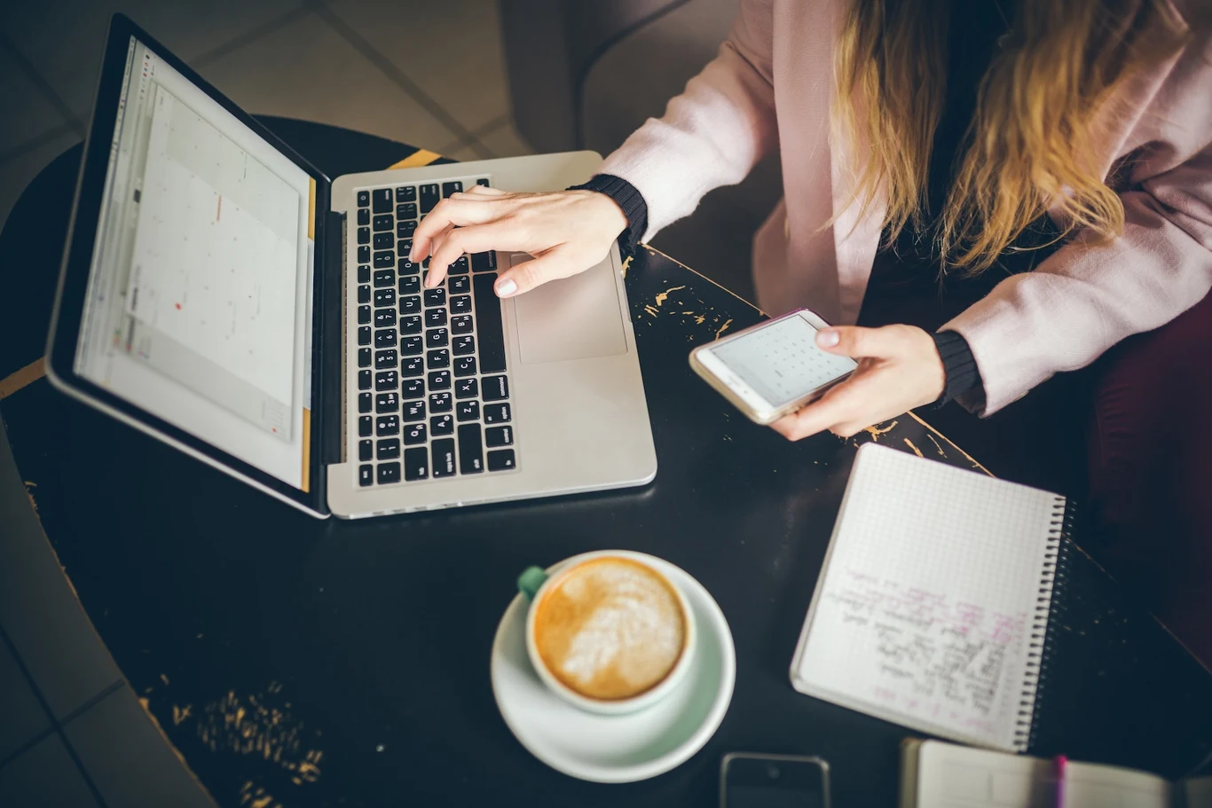 Osoba pracująca przy laptopie, trzymająca telefon w jednej ręce, obok leży notes z zapiskami oraz filiżanka kawy na okrągłym stoliku.