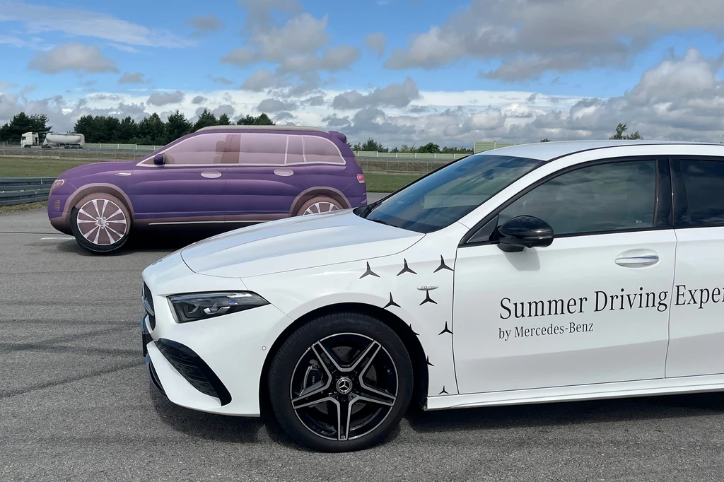 Biała osobowa limuzyna marki Mercedes-Benz z napisem Summer Driving Experience stoi na asfalcie, w tle widoczny prototyp pojazdu w kolorze fioletowym z uproszczonym wyglądem, przypominającym atrapę samochodu testowego.