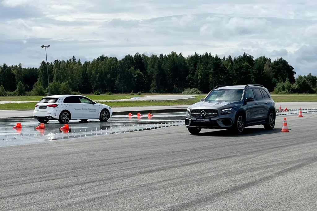 Czarny SUV marki Mercedes oraz biały samochód osobowy jadą po torze testowym z rozstawionymi pachołkami i mokrą nawierzchnią, w tle drzewa i niebo z chmurami.