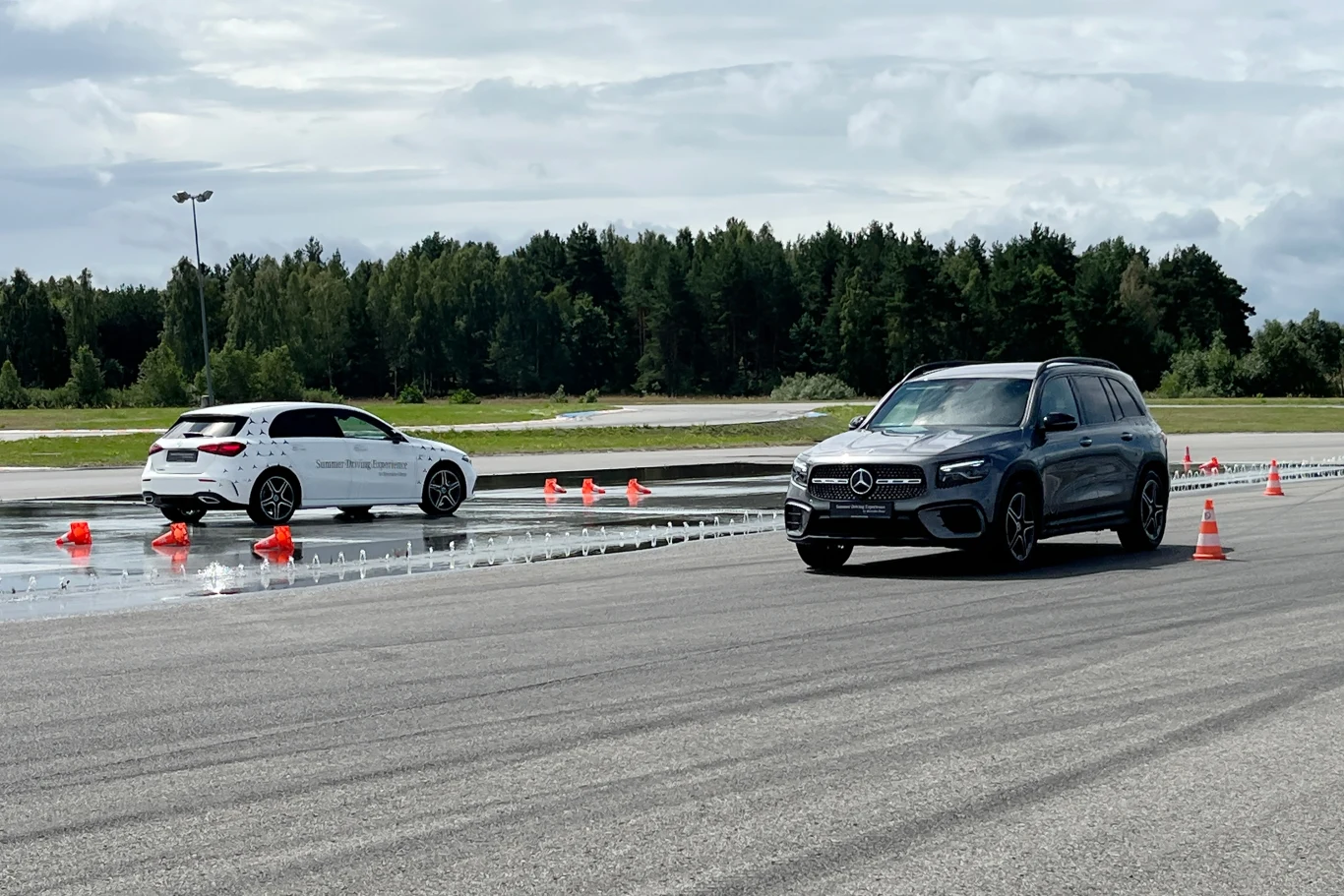 Czarny SUV marki Mercedes oraz biały samochód osobowy jadą po torze testowym z rozstawionymi pachołkami i mokrą nawierzchnią, w tle drzewa i niebo z chmurami.