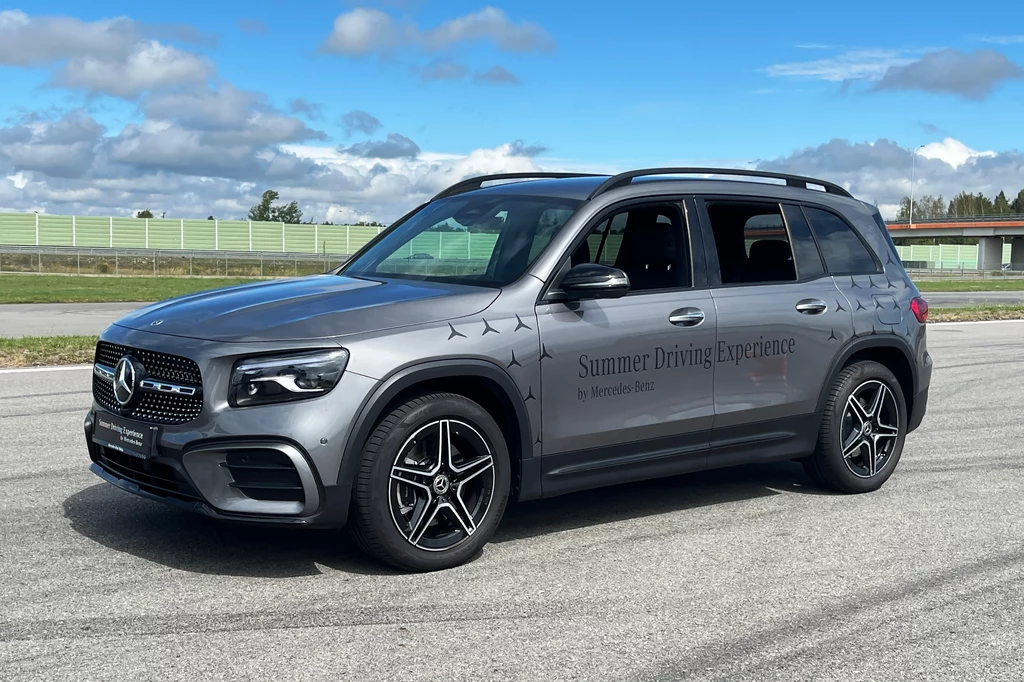 Srebrny SUV marki Mercedes-Benz zaparkowany na szerokim, pustym asfalcie, po bokach toru widoczna niska bariera ochronna i rozległy trawiasty teren z drzewami w oddali, na aucie napis Summer Driving Experience.