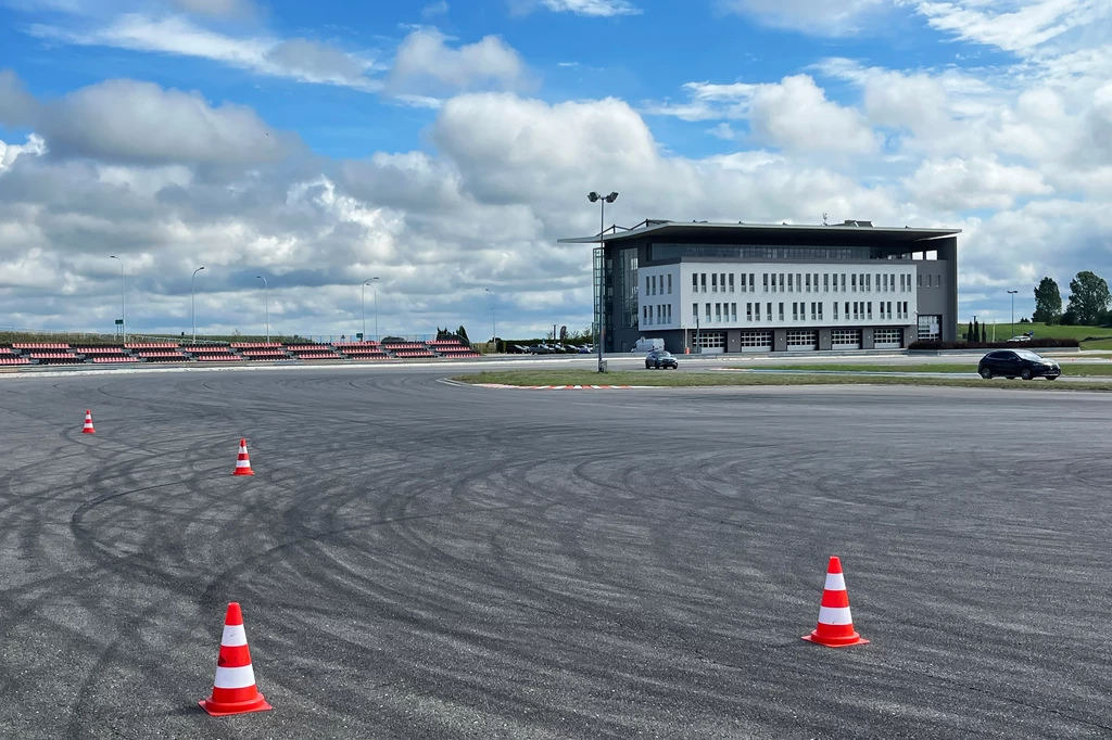 Rozległy asfaltowy tor, na którym ustawiono pomarańczowo-białe pachołki, w tle nowoczesny budynek oraz dwa samochody, całość pod niebieskim niebem z chmurami.