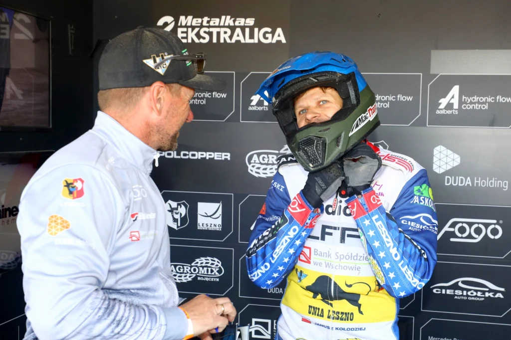 Janusz Kołodziej w rozmowie z trenerem Rafałem Okoniewskim Dwóch mężczyzn podczas rozmowy, jeden ubrany w strój żużlowca i kask, stojący na tle tablicy z logotypami sponsorów.