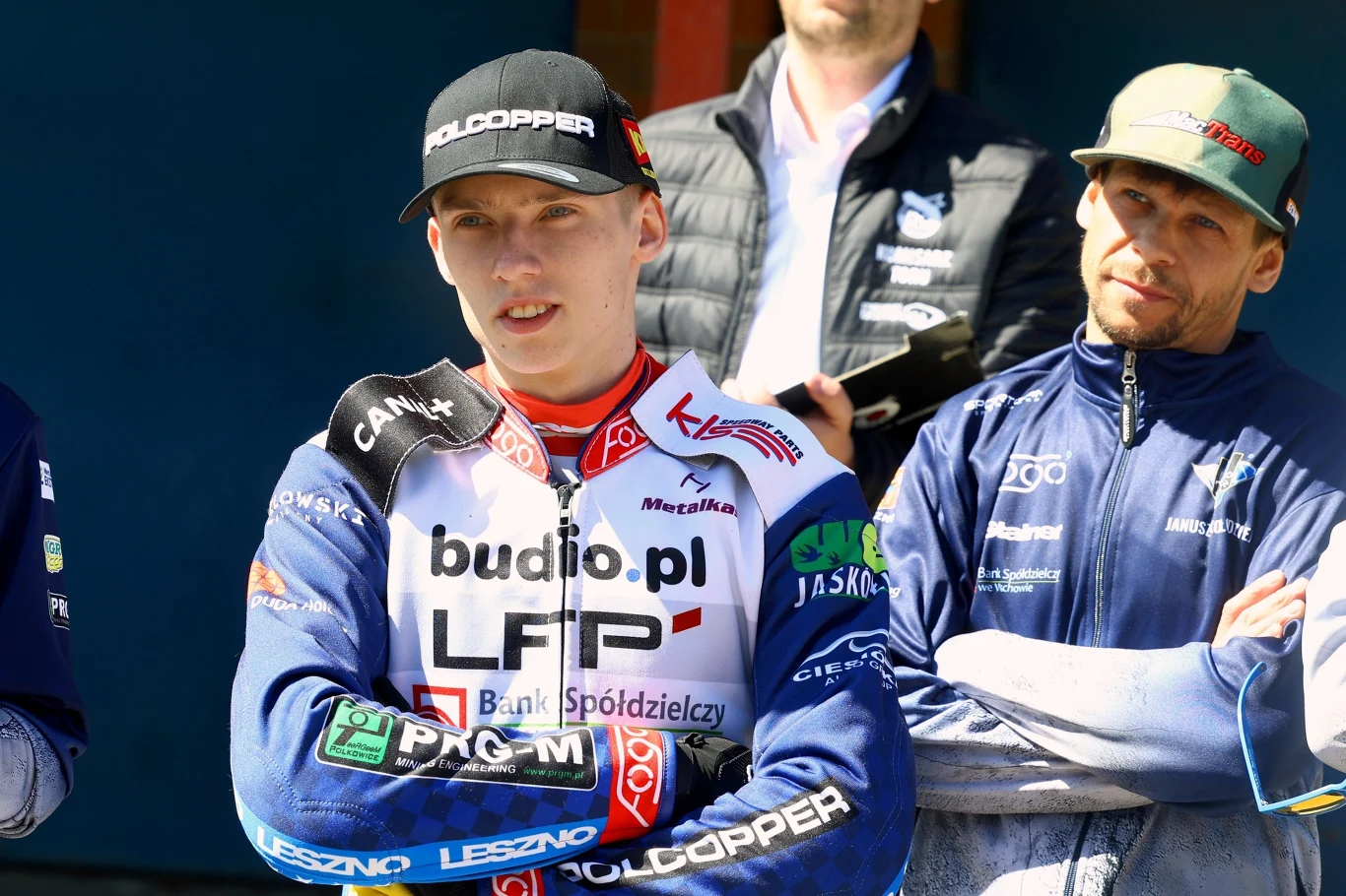 Młody żużlowiec w profesjonalnym stroju z licznymi sponsorami na kurtce stoi z założonymi rękami, obok drugi mężczyzna w sportowej bluzie i czapce, za nimi widoczny inny mężczyzna, wszyscy w świetle słonecznym.