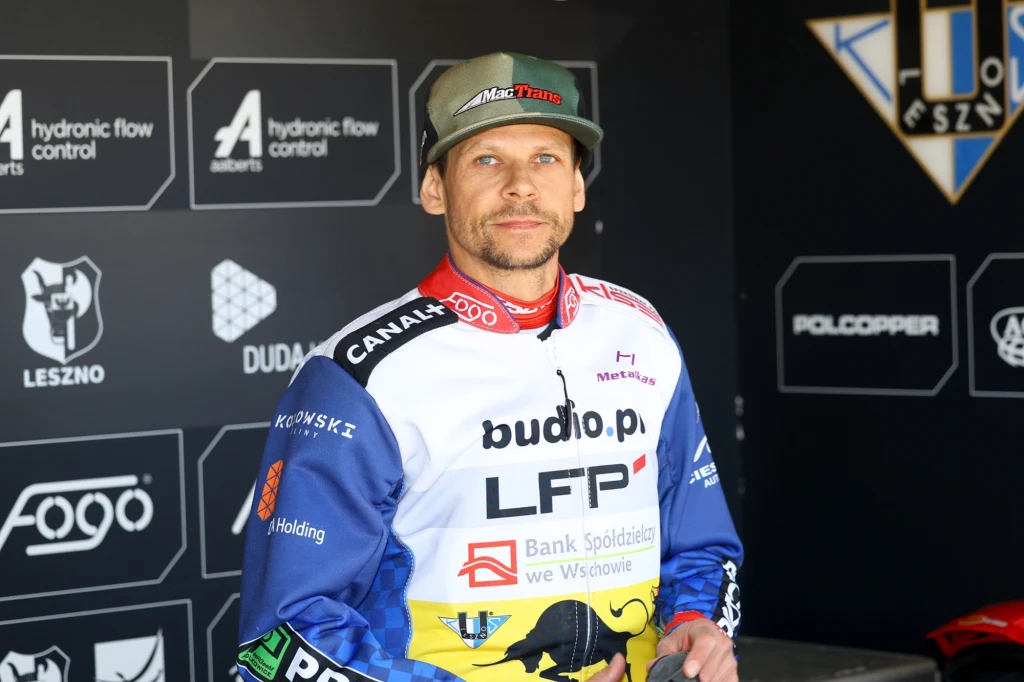 Janusz Kołodziej, kapitan FOGO Unii Leszno Mężczyzna w stroju sportowym z licznymi logotypami sponsorów, stojący na tle ścianki z logo klubu sportowego oraz firm. Ubrany w czapkę z daszkiem i kombinezon motocrossowy, patrzy prosto w obiektyw.