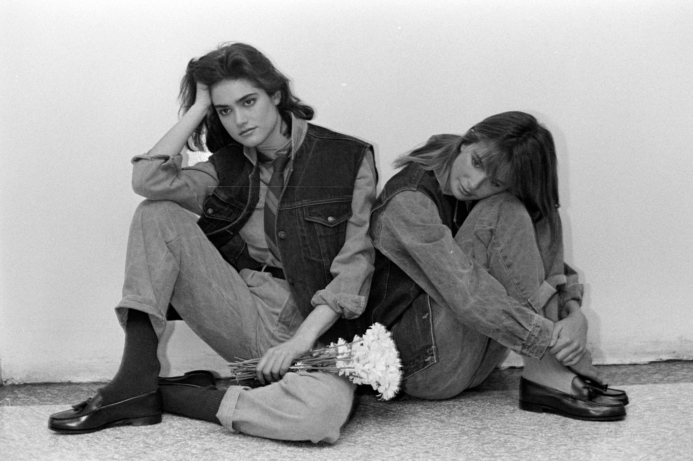 Calvin Klein Jeans 1983 Dwie młode kobiety siedzą oparte plecami o siebie na podłodze, ubrane w dżinsowe ubrania i kamizelki, jedna z nich trzyma białe goździki, mają poważne miny.