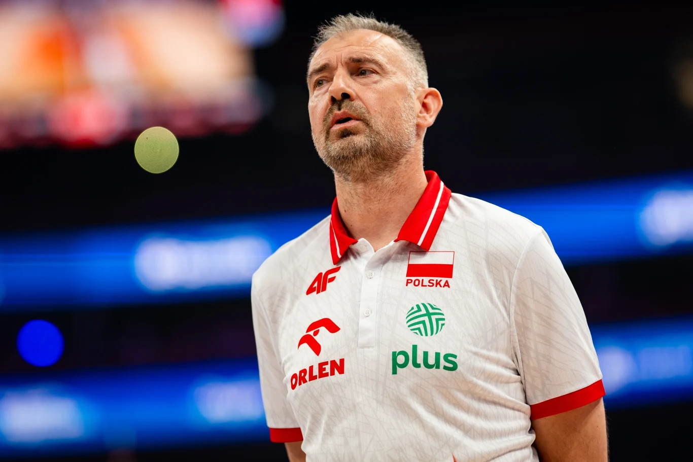 Nikola Grbić Trener w biało-czerwonej koszulce z logotypami sponsorów, stojący na tle rozmytego stadionu sportowego, wyraźnie skupiony, z napisem "Polska" na piersi.