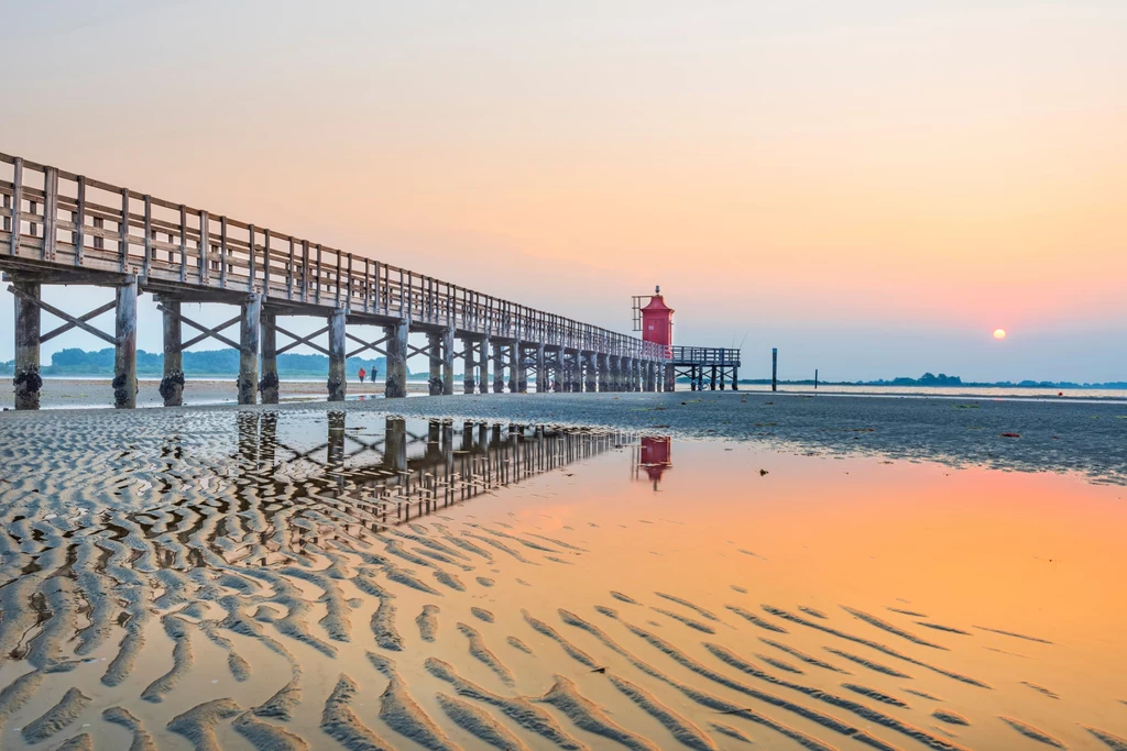 Plaże w Lignano Sabbiadoro charakteryzują się malowniczą linię brzegową długi drewniany pomost prowadzący do czerwonej latarni morskiej, odbijającej się w spokojnej wodzie podczas zachodu słońca, z widocznymi piaszczystymi żłobieniami na pierwszym planie