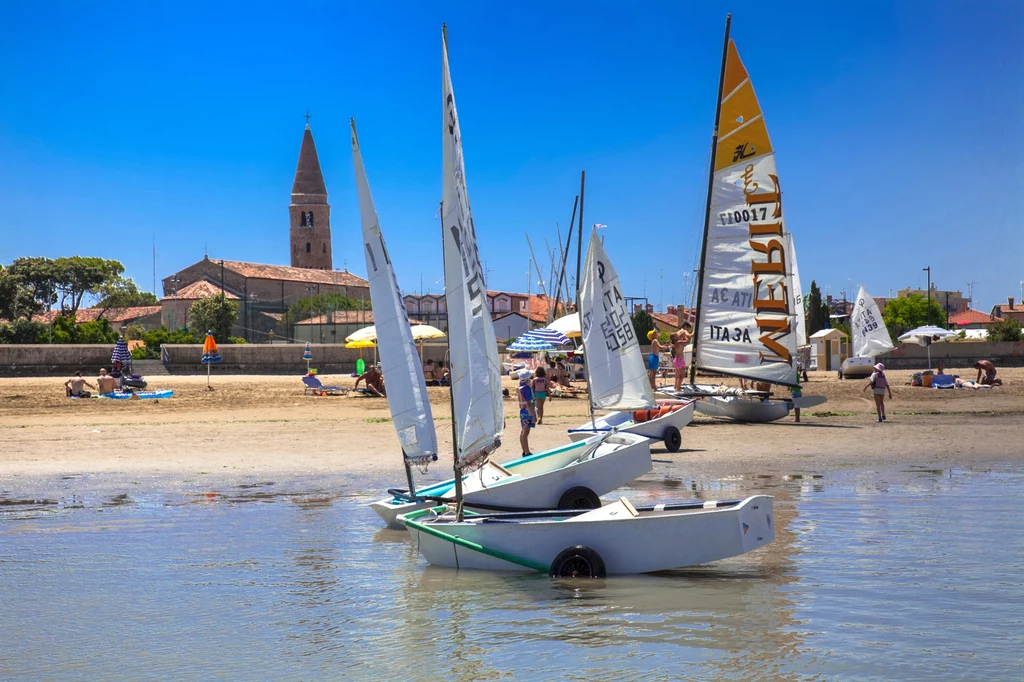 W Caorle można podziwiać nie tylko czyste plaże ale i zdumiewające zakątki historycznego miasta Małe żaglówki ustawione przy brzegu spokojnej plaży, w tle widoczna zabudowa nadmorskiej miejscowości z charakterystyczną wieżą kościoła, a na plaży wypoczywający ludzie pod kolorowymi parasolami.