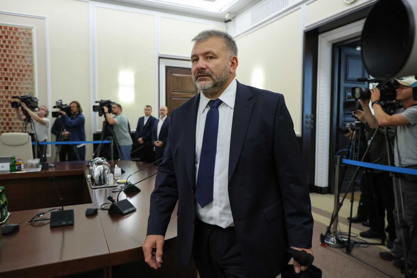 Minister sprawiedliwości Waldemar Żurek Mężczyzna w garniturze i krawacie wchodzi do eleganckiej sali konferencyjnej, w której znajdują się kamery, dziennikarze oraz inni uczestnicy spotkania.