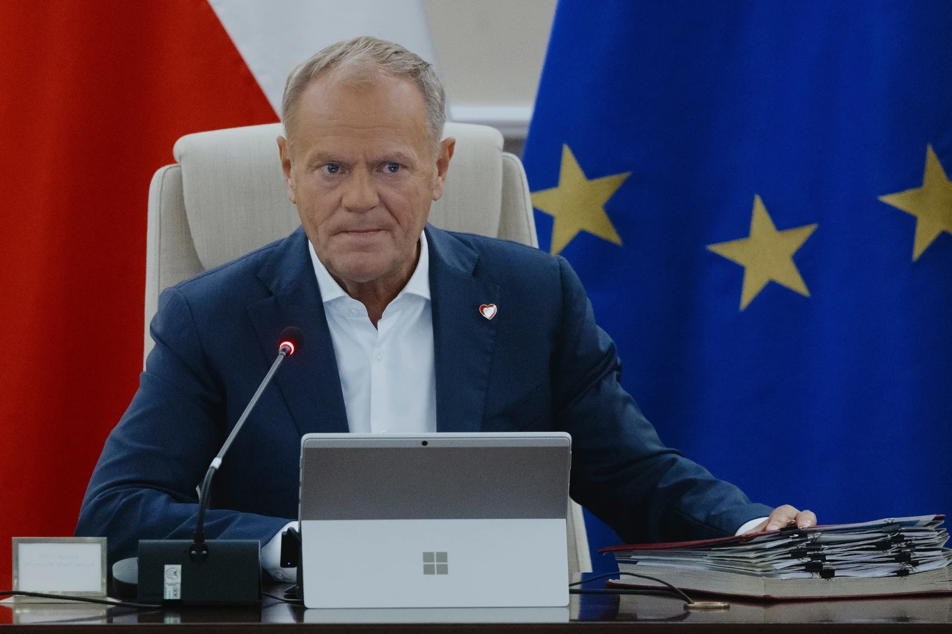 Premier Donald Tusk Mężczyzna w garniturze siedzący przy stole konferencyjnym, przed nim laptop i mikrofon, w tle widoczne fragmenty flag Polski i Unii Europejskiej oraz stos dokumentów.