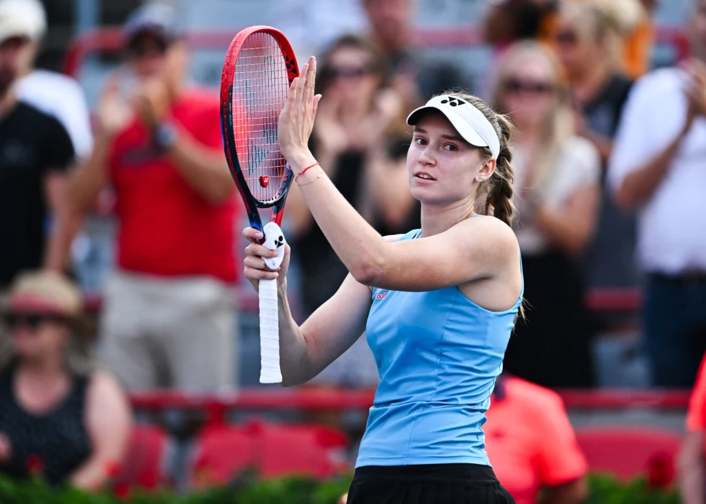 Tenisistka w jasnoniebieskiej koszulce i białej czapce trzymająca rakietę do tenisa, klaszcząca dłonią o rączkę rakiety, w tle widoczna publiczność ożywiona oglądanym meczem.