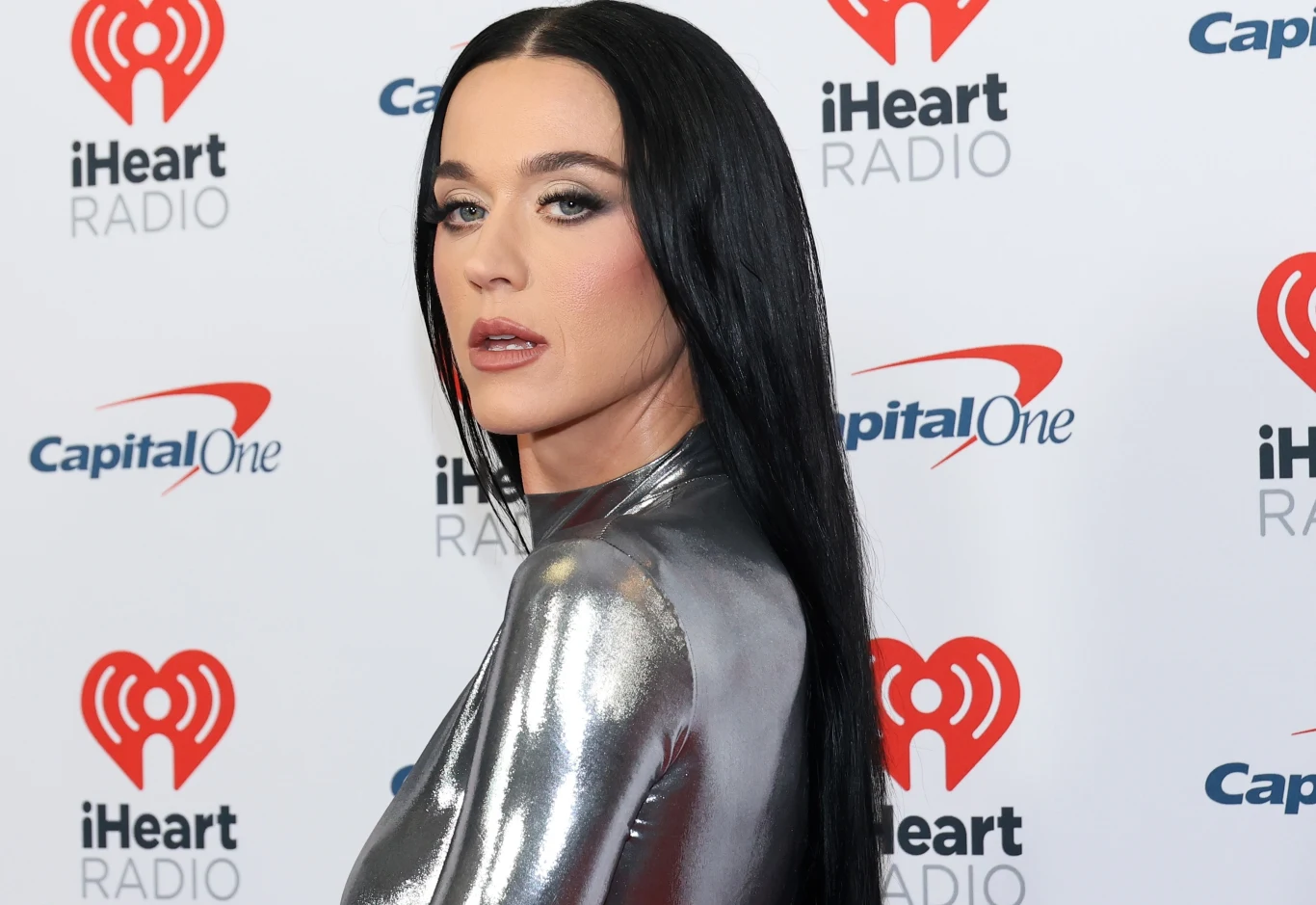 Fanka Katy Perry zemdlała na scenie Kobieta w dopasowanej, srebrnej kreacji z długimi czarnymi włosami pozuje na tle ściany z logo iHeartRadio oraz Capital One.