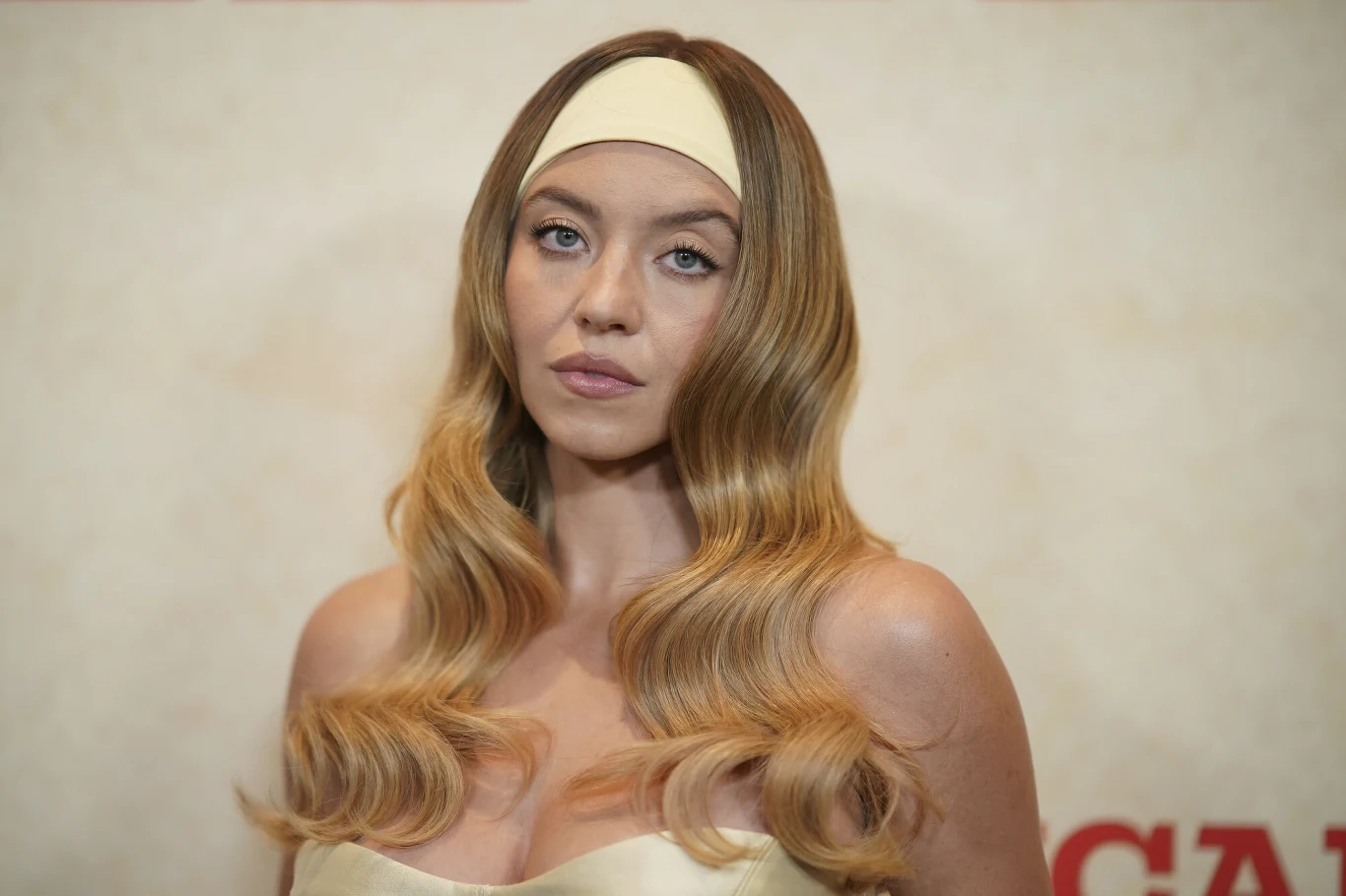 Sydney Sweeney przykuła wzrok swoją stylizacją Kobieta z długimi, falowanymi blond włosami i opaską na głowie, ubrana w jasny, elegancki strój, patrzy prosto w obiektyw na neutralnym tle.