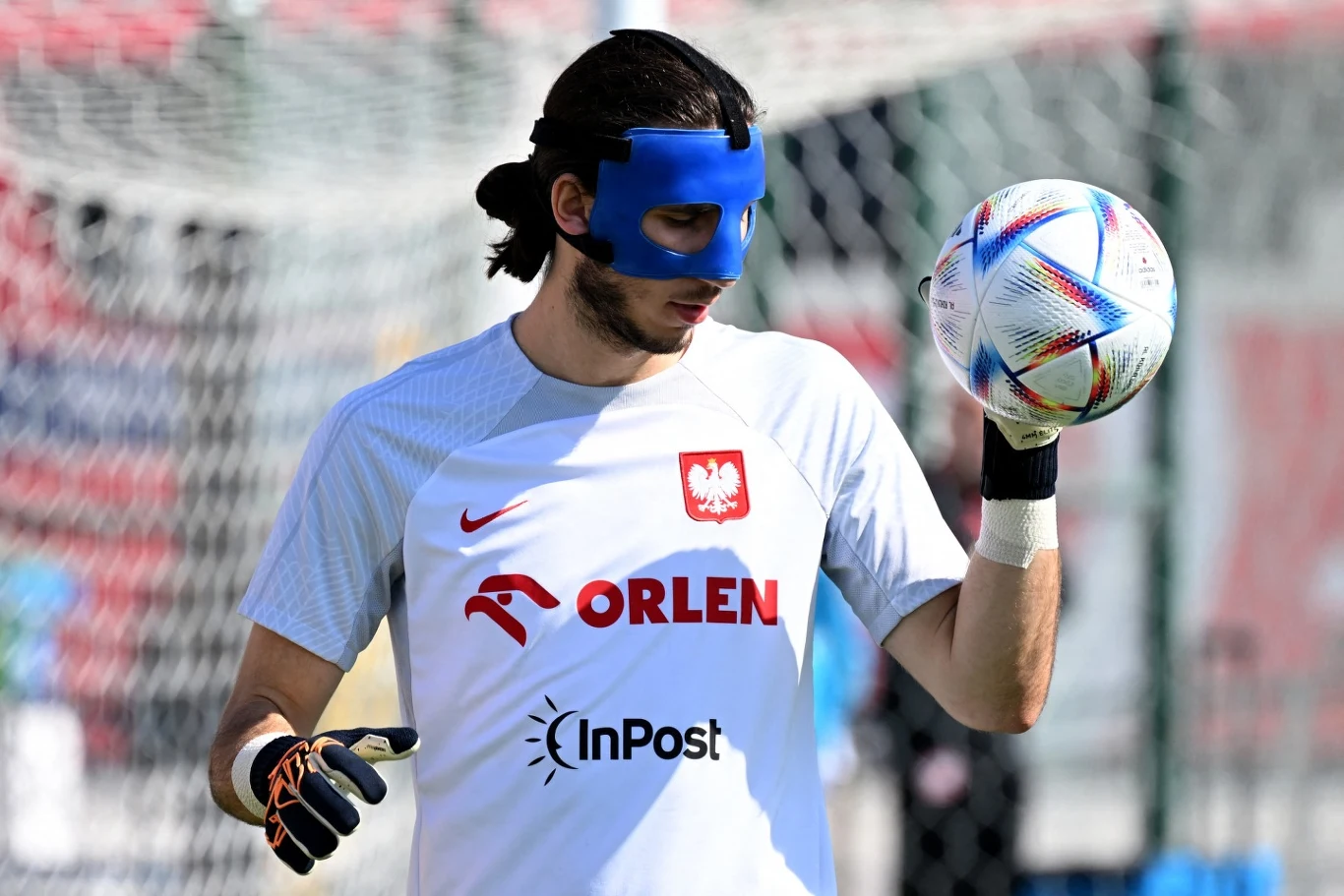 Bramkarz w białej koszulce Polski z logotypami sponsorów trzyma w ręku piłkę nożną, na twarzy ma niebieską maskę ochronną, w tle bramka i piłkarskie boisko