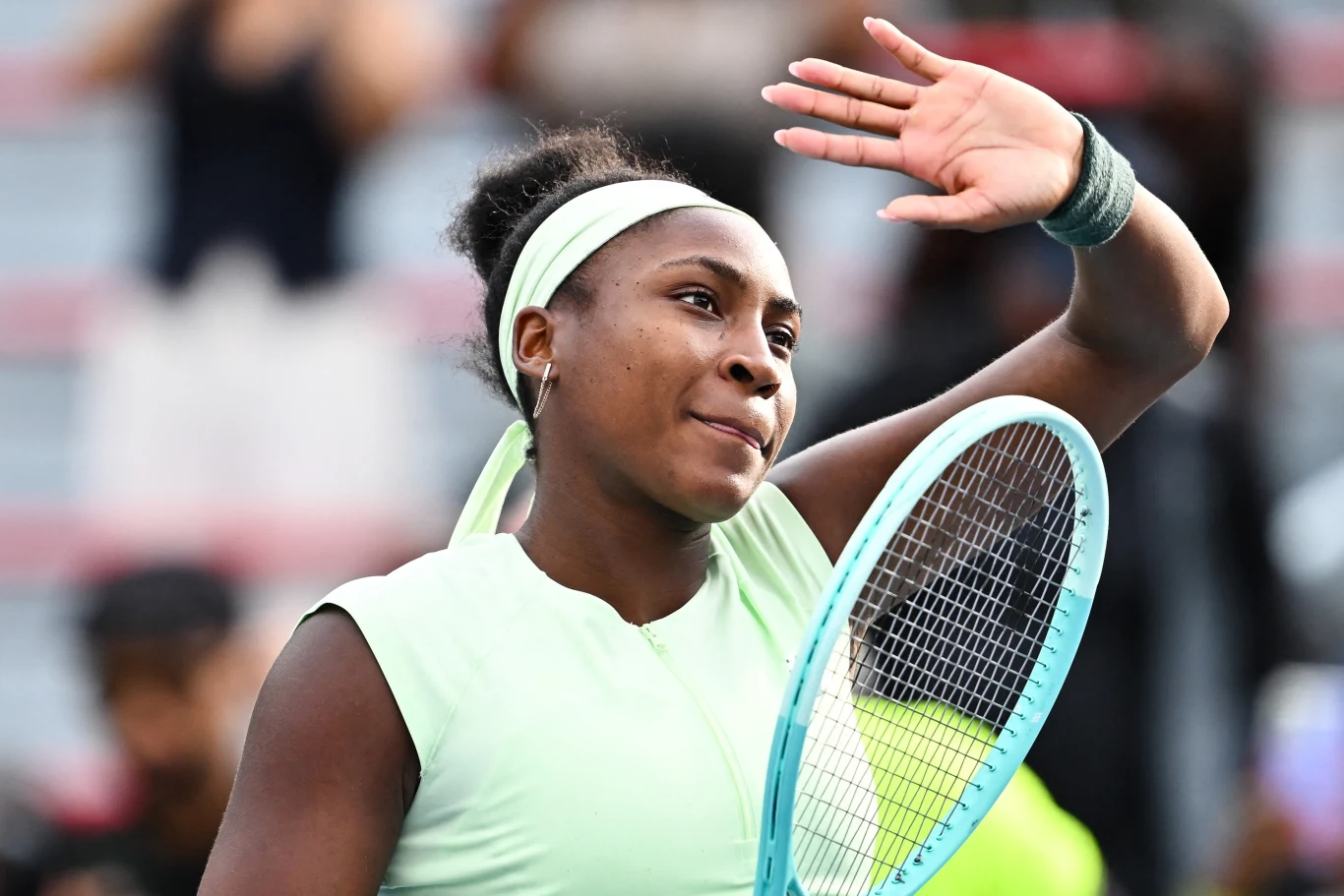 Młoda tenisistka w zielonym stroju trzymająca rakietę do tenisa i machająca ręką, delikatny uśmiech na twarzy, w tle rozmyte sylwetki widzów.