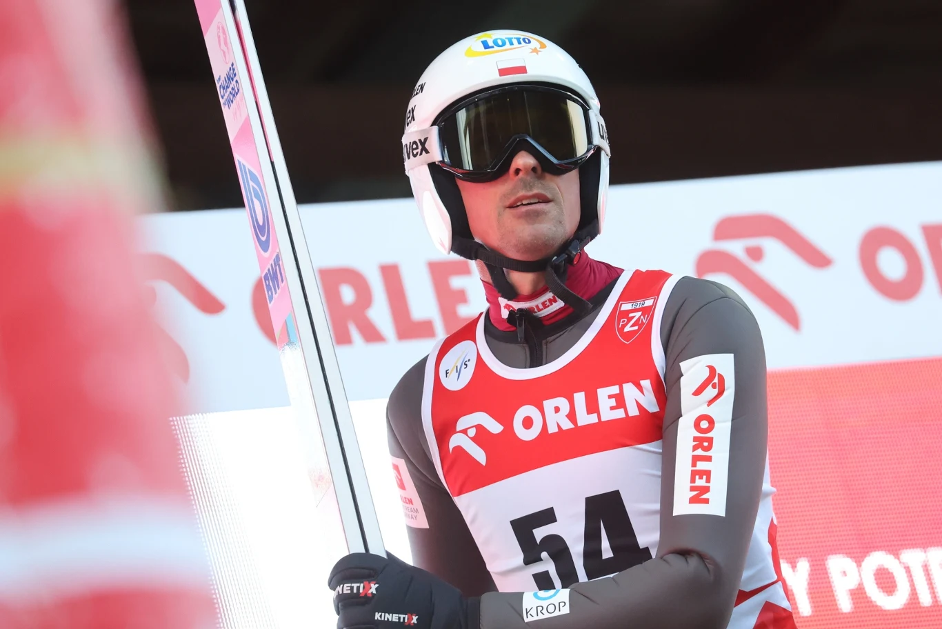 Piotr Żyła Zawodnik w kombinezonie narciarskim oraz kasku, trzymający narty na tle tablic sponsorów podczas konkursu skoków narciarskich.